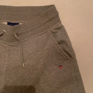 Grå mjukisbyxor från Gant - Snygga grå mjukisbyxor från Gant med snörning i midjan och ribbade muddar. Byxorna har fickor och en liten broderad logotyp på sidan. Perfekta för en avslappnad stil.