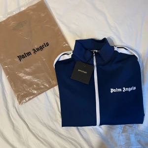 Blå träningsjacka från Palm Angels - Snygg blå träningsjacka från Palm Angels med vita detaljer och dragkedja. Jackan har långa ärmar och en klassisk sportig look med vita ränder längs ärmarna. Perfekt för en stilren och avslappnad stil. Samt med snygg byxorna från Palm angels. 