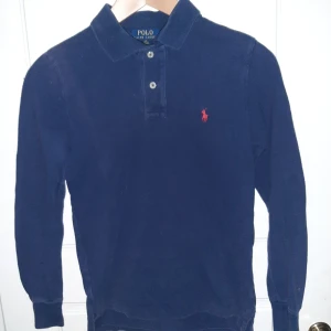 Mörkblå pikétröja från Ralph Lauren - Säljer en klassisk mörkblå pikétröja från Ralph Lauren med den ikoniska röda loggan på bröstet. Tröjan har en krage med knappar och är perfekt för en stilren look. Passar både till vardags och mer uppklädda tillfällen.