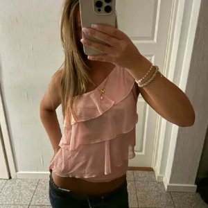 Rosa volanglinne - Säljer ett sött rosa linne med volangdetaljer. Toppen är ärmlös och har en luftig passform, perfekt för varma dagar. Passar bra till både jeans och kjol.