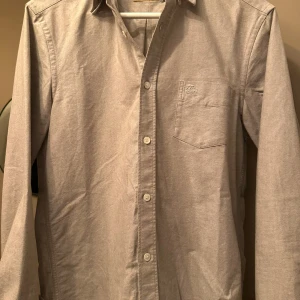 Silver/grå skjorta från Burberry - Snygg silver/grå skjorta från Burberry med klassiskt rutigt mönster på insidan av kragen och ärmsluten. Skjortan har långa ärmar och knappar framtill samt en bröstficka. Perfekt för en stilren look. Den är köpt på NK för längesedan men har knappt användt den.