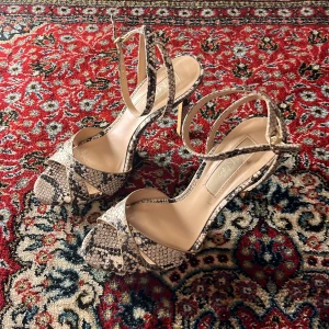 Ormskinnsmönstrade stiletter  - Snygga stiletter med ormskinnsmönster och smala remmar. De har en elegant klack och en öppen tådesign, perfekt för att lyfta din stil. Passar utmärkt till både vardag och festliga tillfällen.