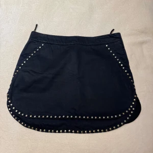 Svart kjol med nitar - Snygg svart kortkjol dekorerad med silverfärgade nitar längs kanterna. Kjolen har en dragkedja på sidan för enkel på- och avtagning. Perfekt för en edgy look!