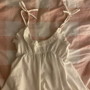 Vit topp med knytband - Söt vit topp med knytband på axlarna och volangdetaljer vid halsringningen. Köpt här på Plick men tyvärr står inte märket eller storlek, skulle dock säga att den är i storlek XS. Toppen är i bra skick och har inga defekter💕