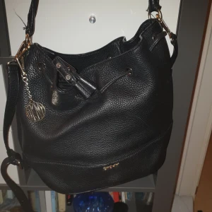 Svart axelväska i skinn - Snygg svart axelväska i skinn med dragkedja och dekorativt hänge. Perfekt för att bära dina dagliga nödvändigheter med stil. Väskan har justerbar axelrem och flera fack för enkel organisering.