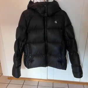 Svart vinterjacka - Svart vinterjacka från Calvin Clein. Använd 1 vinter. Tog storlek M för att kunna ha tjocktröjor under så passar både som M och S. Enda defekten är på baksidan av jackan men syns ej mycket i verkligheten, se sista bilden. Avtagbar huva. Nypris 2200kr