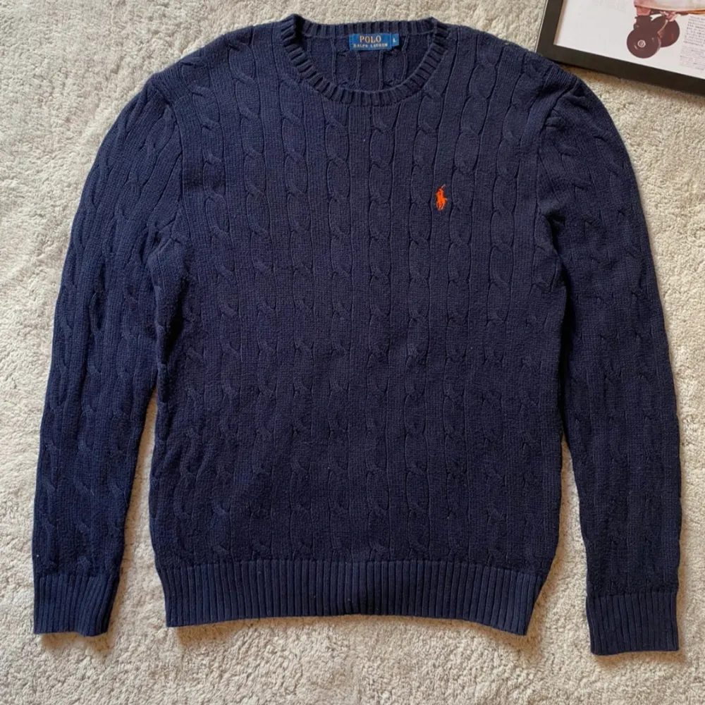 Säljer en snygg mörkblå stickad tröja från Polo Ralph Lauren. Den har ett klassiskt kabelstickat mönster och en broderad orange logga på bröstet. Tröjan är långärmad och perfekt för både höst och vinter. Superbra skick och riktigt stilren!. Tröjor & Koftor.