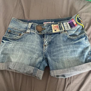 Jeansshorts från Fabu - Säljer ett par snygga jeansshorts från Fabu. De är i en klassisk blå denimfärg med coola detaljer som uppvikta ben och flera patchar. Perfekta för sommaren och har en avslappnad, casual stil. Passar perfekt till en dag på stranden eller en sommarfest!