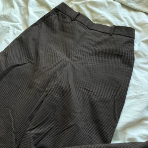 Bruna kostymbyxor  - Bruna kostymbyxor med rutigt mönster och mjukt tyg. De är vida i benen och fullånga. De har fickor på sidorna och resor i midjan. Inga knappar eller dragkedja.