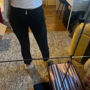 Svarta byxor med knappar - Säljer ett par svarta kostymbyxor byxor med lågmidja och två dekorativa knappar framtill. Byxorna har en normal passform och är perfekta för både vardag och fest. De är bekväma och stilrena, vilket gör dem till ett mångsidigt tillskott i garderoben. Den har en jätte fin och elegant söm på framsidan och fickor på varsin sida.