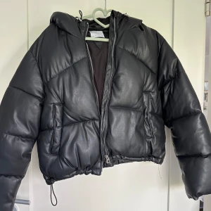 Pufferjacka dam stl S - Svart pufferjacka med luva i skinnimitation storlek S från Bershka. Dragkedja som stängning, öppna fickor. Dragsko i midjan och luvan. Inga defekter, använd fåtal gånger. Är i mycket bra skick.
