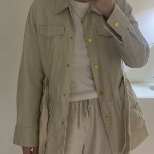 Beige jacka från Bondelid - Säljer en snygg beige jacka från Bondelid i storlek 36. Jackan har långa ärmar och en stilren design med knappar och ett knytbälte i midjan för en fin passform. Perfekt för vår och höst!