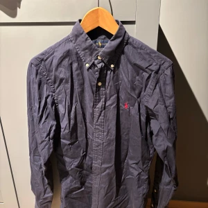 Mörkblå skjorta från Ralph Lauren - Säljer en snygg mörkblå skjorta från Ralph Lauren i bra skick. Skjortan har långa ärmar och klassiska knappar framtill. Den är perfekt för både vardag och fest, med en liten röd broderad logga på bröstet som ger en stilren touch. Passar perfekt till jeans eller chinos!