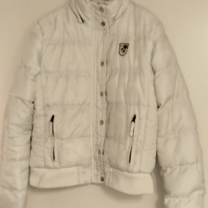 Vit dunjacka med emblem - Säljer en snygg vit dunjacka med ett coolt emblem på bröstet. Jackan har en hög krage och stängs med knappar. Perfekt för kyliga dagar och håller dig varm och stilren. Den har långa ärmar och praktiska fickor med dragkedja. Passar perfekt till både vardag och fest!