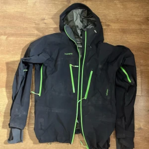Svart vindjacka från Norrøna - Säljer en svart vindjacka från Norrøna med gröna dragkedjor. Jackan har en justerbar huva och flera praktiska fickor. Perfekt för utomhusaktiviteter och håller dig skyddad mot vinden. Passar bra för höst och vår.