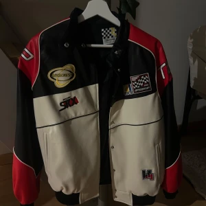 Röd och svart racingjacka - Säljer en cool racingjacka i rött, svart och vitt med flera tryck och loggor. Jackan har en sportig stil med knappar och ribbade muddar. Perfekt för dig som gillar motorsport och vill ha en unik look!