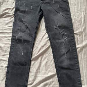 Säljer ett par svarta jeans från Dsquared2 i bra skick. De har en snygg slitningseffekt på framsidan och knappar med märkets logga. Perfekta för en casual look eller en utekväll. Passformen är slim och de är tillverkade i ett bekvämt denimtyg.