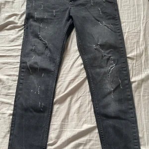 Svarta jeans från Dsquared2 - Säljer ett par svarta jeans från Dsquared2 i bra skick. De har en snygg slitningseffekt på framsidan och knappar med märkets logga. Perfekta för en casual look eller en utekväll. Passformen är slim och de är tillverkade i ett bekvämt denimtyg.