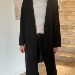 Svart kappa med bälte - Säljer en elegant svart kappa i mycket bra skick. Den har långa ärmar och ett bälte i midjan för en snygg passform. Perfekt för höst och vinter, och funkar lika bra till vardags som till fest. Kappan har en mjuk krage som ger extra stilpoäng. Passa på att uppgradera din garderob med denna klassiska pjäs!
