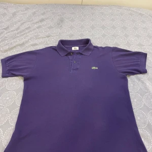 Lila pikétröja från Lacoste - Säljer en snygg lila pikétröja från Lacoste i bra skick. Den har klassisk krage med två knappar och den ikoniska krokodilloggan på bröstet. Perfekt för både vardag och lite finare tillfällen. Passar bra till jeans eller chinos!