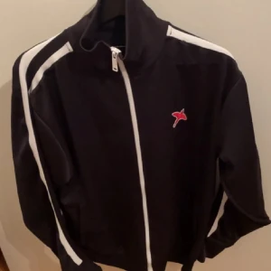 Svart jacka från Axel Arigato - Säljer en snygg svart tröja från Axel Arigato med vita detaljer och dragkedja. Den har en röd fågel logga på bröstet och vita ränder längs ärmarna. 