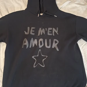 Svart hoodie från Gina Tricot - Säljer en svart hoodie från Gina Tricot med texten 'JE M'EN AMOUR' och en stjärna på framsidan. Den har en mysig huva med dragsko och är perfekt för en avslappnad stil. Passar bra till både vardag och chillkvällar. 🖤
