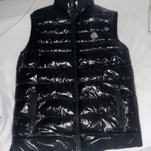 Säljer en snygg svart dunväst från Moncler i glansigt material. Västen har en dragkedja framtill och Moncler-logga på bröstet. Perfekt för höst och vinter, och den är i mycket bra skick. Passar både till vardags och festliga tillfällen. Västen har tecken på användning men inget som går att märka i helheten där av priset :) Passar fint till både tjejer & killar 🥰