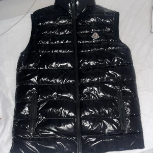 Svart dunväst från Moncler - Säljer en snygg svart dunväst från Moncler i glansigt material. Västen har en dragkedja framtill och Moncler-logga på bröstet. Perfekt för höst och vinter, och den är i mycket bra skick. QR koden fungerar perfekt. Passar både till vardags och festliga tillfällen. Västen har tecken på användning men inget som går att märka i helheten där av priset :) Passar fint till både tjejer & killar 🥰