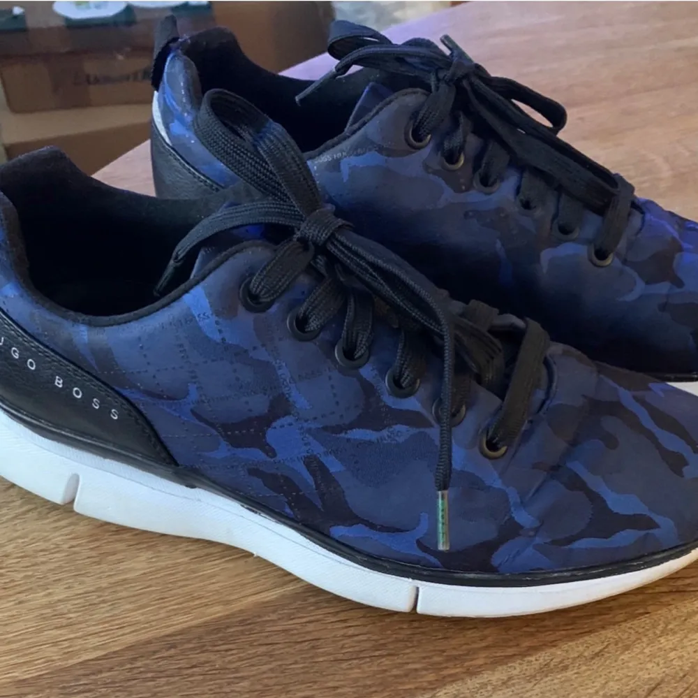 Snygga blå sneakers från Hugo Boss med ett coolt camouflage-mönster. Skorna har svarta snören och en vit sula som ger en sportig look. Perfekta för både vardag och lite mer avslappnade tillfällen. Märket syns tydligt på sidan av skorna.Dessa är äkta och köpt i lokal butik här i Malmö. Det är boss grön serie. Det syns på skosnöre. Äkta på alla vis. Använda men syns knapp så i nyskick!  St 39 och 25,3 cm med skosticka i innermått. Syns enbart lite på sulan att dom är använda. Nypris ca 1500:- . Skor.