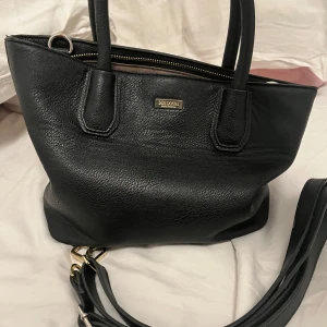 Svart handväska från Don Donna - Snygg svart handväska från Don Donna i skinn. Den har en praktisk dragkedja och avtagbar axelrem. Den rymmer otroligt mycket trots dess storlek. Sista bilden är till referens av storleken på väskan! Fläckar insidan av väskan. Köpt för ca 300kr 