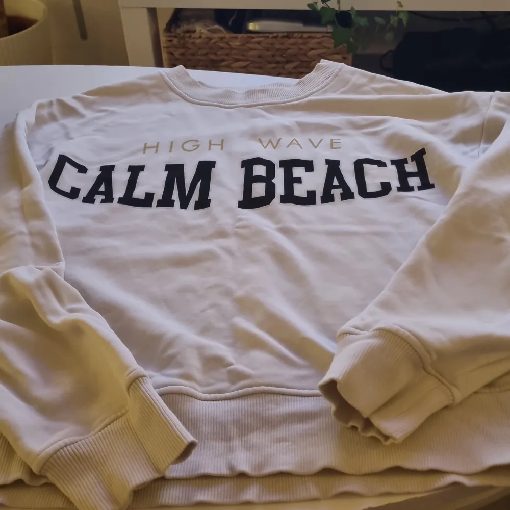 Säljer en supermysig vit sweatshirt från Lindex i storlek XS. Den har texten 'High Wave Calm Beach' tryckt på framsidan i svart och guld. Perfekt för en avslappnad dag eller en promenad på stranden. Tröjan har långa ärmar och ribbade muddar vid ärmslut och nederkant.. Tröjor & Koftor.