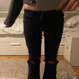 Svarta jeans med slitningar - Säljer ett par svarta jeans med coola slitningar på knäna. De är i bootcut-stil och är lowwaist!! Jätte snygga!!! Vill ha kvar men de är tyvärr för korta🥹🥹. Ja har gjort hålet själv! 