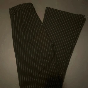 Svarta randiga kostymbyxor från Gina Tricot - Snygga svarta kostymbyxor från Gina Tricot med vita ränder. Perfekta för både kontoret och festliga tillfällen. Byxorna har en klassisk passform med hög midja och raka ben. De är i nytt skick och har en stilren design som passar till det mesta. Ett måste i garderoben för en chic look!