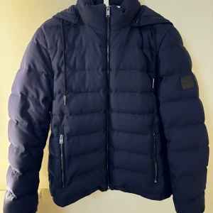 Mörkblå pufferjacka från Boss - Säljer en snygg mörkblå pufferjacka från Boss. Jackan är i mycket bra skick och har en quiltad design med dragkedja framtill. Den är perfekt för höst och vinter med sitt Green Storm System-tyg från Loro Piana som skyddar mot regn och vind. Storleken är inte specificerad, men den ser ut att ha en normal passform. Perfekt för att hålla sig varm och stilren!