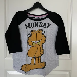Grå och svart Garfield-tröja med trekvartsärm - Säljer en superkul grå och svart tröja med Garfield-tryck och texten 'Monday' på framsidan. Tröjan har trekvartsärmar och är perfekt för en avslappnad look, i bra skick. Perfekt för alla Garfield-fans!