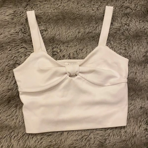 Vit croppad topp med rosett - Säljer en superfin vit croppad topp med breda axelband och en söt rosett framtill. Perfekt för sommardagar eller en utekväll. Toppen är i ett mjukt och bekvämt material som sitter skönt på kroppen. Passar perfekt till både jeans och kjol!