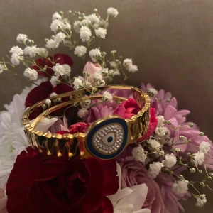 Rostfritt stål armband med öga-design - Snyggt guld färger armband i rostfritt stål med en unik öga-design i mitten. Armbandet har en blå och vit detalj runt ögat som ger en cool kontrast. Perfekt för att ge din outfit en extra touch av stil. Passar både till vardags och festliga tillfällen!