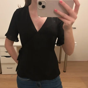 Svart omlottblus - Snygg svart omlottblus med korta ärmar. Perfekt för både vardag och fest. Blusen har en elegant v-ringning och knytning i sidan för en smickrande passform. 