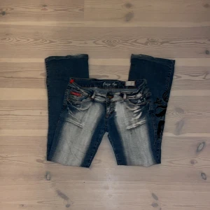 Blå jeans med drakbroderi från Crazy Age - Säljer ett par coola blå jeans från Crazy Age med ett snyggt svart drakbroderi på benet. Det är väldigt lågmidajde och det är boot cut/flare. Jeansen har en sliten look med ljusa partier och är i bra skick.