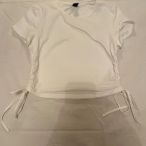 Vit kortärmad topp från Shein - Säljer en snygg vit kortärmad topp från Shein i storlek M. Toppen har knytband på sidorna för en justerbar passform och är perfekt för sommardagar. Materialet är mjukt och bekvämt, vilket gör den idealisk för både vardag och fest. Passa på att uppdatera garderoben med denna stilrena topp!