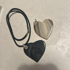 Halsband med hjärtformade stenar - Snyggt halsband med två hjärtformade hängen i olika färger. Ena stenen är i jordnära beige och bruna toner medan den andra är mörkgrå. Båda hängena har en liten metallbricka med initialer. Kedjan är i svart flätat material med ett enkelt lås. Perfekt för att ge en personlig touch till din outfit!