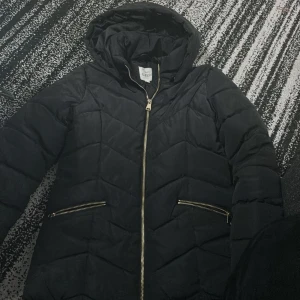 Svart dunjacka från Kappa - Säljer en svart dunjacka från Kappal  i storlek 152. Jackan har en snygg quiltad design med dragkedja och två fickor framtill. Perfekt för kalla vinterdagar med sin varma och bekväma passform. Huvan ger extra skydd mot väder och vind. Passar både till vardags och lite finare tillfällen.