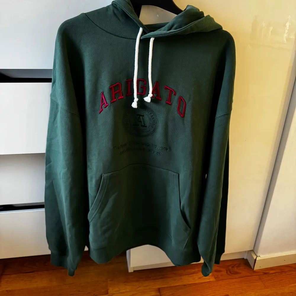 Säljer en snygg grön hoodie från Axel Arigato med röd text på bröstet. Den har en klassisk känguruficka och justerbar huva med vita snören. Perfekt för en avslappnad stil och passar bra till både jeans och joggers. Hör av dig vid intresse!Aldrig använd! Prislapp kvar. Hoodies.