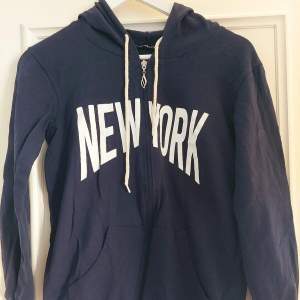 Säljer en mörkblå hoodie med stort vitt 'New York'-tryck på framsidan. Den har en dragkedja och en mysig huva med snören. Perfekt för en avslappnad stil eller en promenad i stan. Stor ficka framtill för att värma händerna eller förvara småsaker.