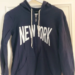 Mörkblå hoodie med New York-tryck - Säljer en mörkblå hoodie med stort vitt 'New York'-tryck på framsidan. Den har en dragkedja och en mysig huva med snören. Perfekt för en avslappnad stil eller en promenad i stan. Stor ficka framtill för att värma händerna eller förvara småsaker.