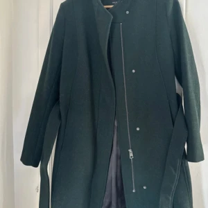 Mörkgrön kappa från Selected Femme - Säljer en elegant mörkgrön kappa från Selected Femme i storlek 36. Den har en stilren design med dragkedja och knappar framtill. Perfekt för höst och vinter, och ger en sofistikerad look. Kappan är i mycket bra skick och har ett bälte för att framhäva midjan.
