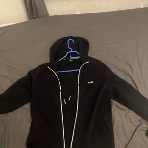 Svart hoodie från Boss - Säljer en snygg svart hoodie från Boss i mycket bra skick. Den har en dragkedja framtill och en luva med dragsko. På bröstet finns en diskret Boss-logga och längs nederkanten är det ett coolt mönster med Boss-text. Perfekt för höst och vår!