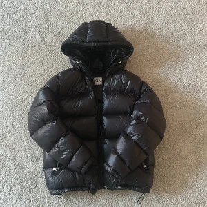 Svart pufferjacka från Zara - Säljer en snygg svart pufferjacka från Zara. Den är perfekt för kalla dagar med sin tjocka vaddering och har en praktisk huva. Jackan har dragkedja framtill och justerbara snören i nederkant för bästa passform. Perfekt för vintern! 🖤