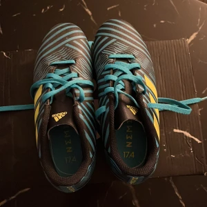 Adidas Nemeziz 17.4 fotbollsskor - Oanvända snygga Adidas Nemeziz 17.4 fotbollsskor i svart och blått med gula detaljer. Skorna har ett randigt mönster och snörning för optimal passform. Perfekta för fotbollsplanen med sina greppvänliga dobbar. Perfekt för barn som gillar att lira fotboll.