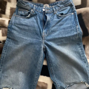 Blå jeans med slitningar - Säljer ett par blå jeans med slitningar på knäna. De har en klassisk femficksdesign och en dragkedja med knapp framtill. Perfekta för en avslappnad stil. Högmidjade/midwaist